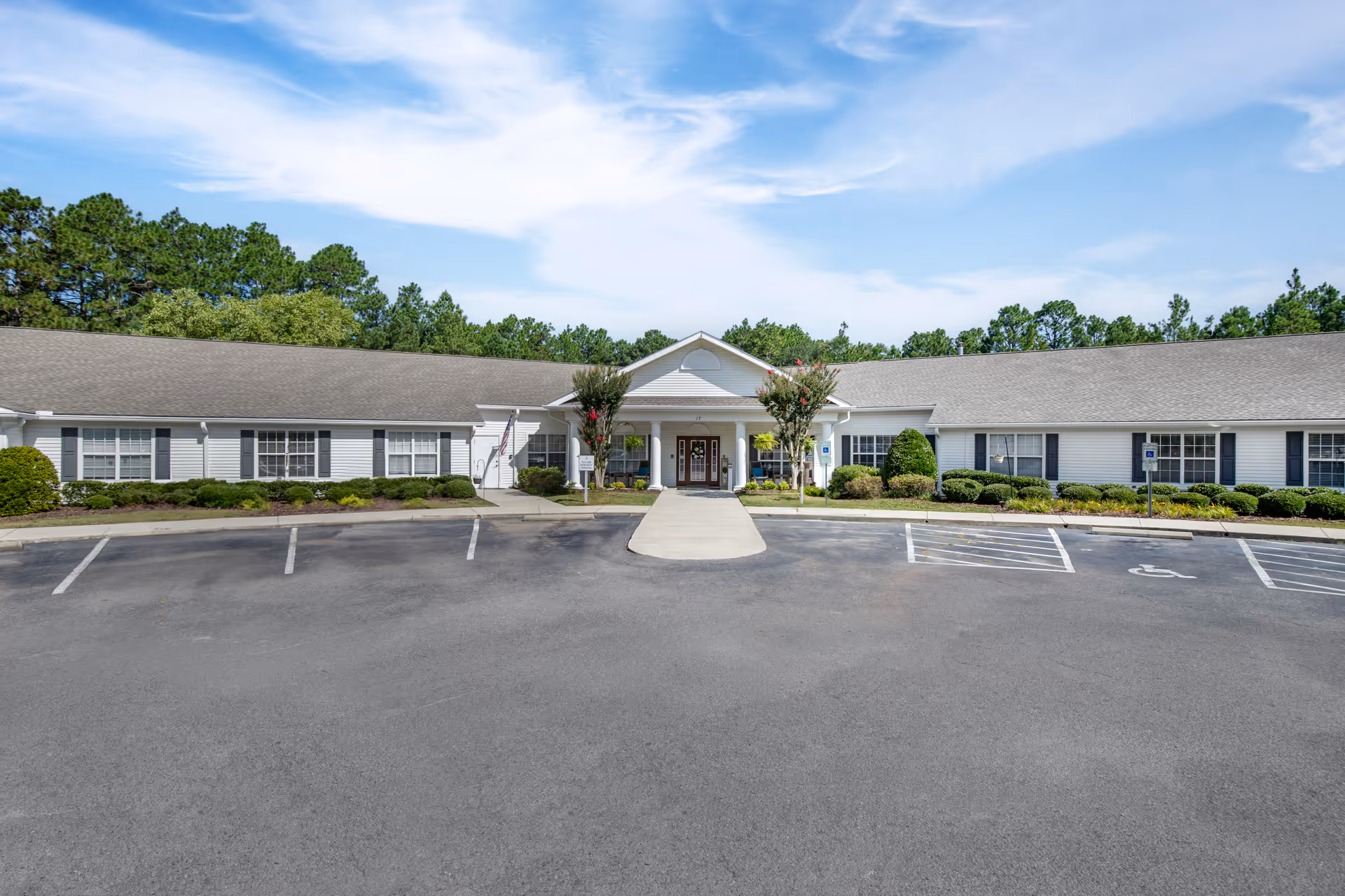 Front exterior view of a single-story senior living facility building with white siding, a gray shingled roof, and a central entrance with columns. There are landscaped bushes and trees around the building, a paved parking lot with marked spaces including handicapped spots, and a clear blue sky with some clouds above.