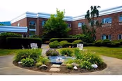 Outdoor garden area with a small circular flower bed featuring a water fountain surrounded by plants and rocks. Two white benches are placed on the paved walkway around the garden. The background shows a two-story brick building with windows and some trees and shrubs.