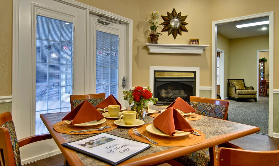 A dining table set for four with folded rust-colored napkins, yellow cups and saucers, and a vase of red roses in the center. The table is in a cozy room with a fireplace, a sunburst mirror above the mantel, and French doors with blinds leading outside. A menu titled 'Taste Of Home Cafe' is on the table. In the background, there is a hallway with a chair and another room visible.
