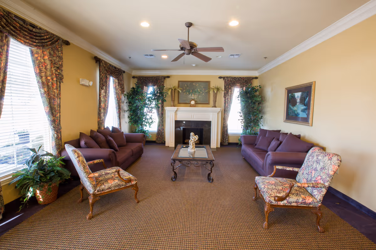A cozy living room with two purple sofas facing each other, two floral upholstered armchairs, a glass coffee table with a small statue, a white fireplace with decorative plants and a clock on the mantel, floral curtains on the windows, and framed artwork on the walls.