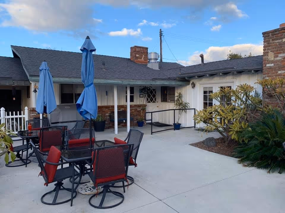 Outdoor patio area at Family Choice Senior Living with several glass-top tables and red cushioned chairs. Blue umbrellas are closed and positioned over the tables. The patio is adjacent to a single-story building with white siding and brick accents. There are potted plants and some greenery around the patio area under a partly cloudy sky.
