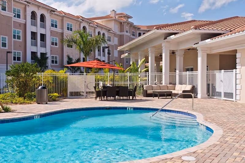 Outdoor swimming pool area with clear blue water, surrounded by a brick-paved deck. There is a seating area with a beige cushioned sectional sofa under a covered patio with white columns. Nearby, there is a dining table with black chairs and a large red umbrella. In the background, there is a multi-story building with pinkish walls and multiple windows, along with palm trees and other greenery.