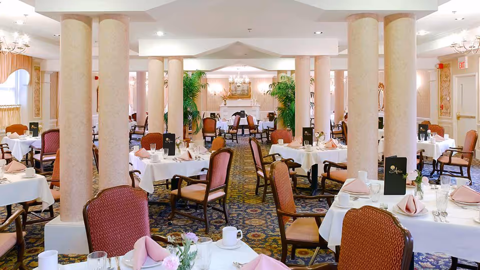 Formal dining room with multiple tables set with white linens and pink napkins, upholstered chairs, and decorative columns.