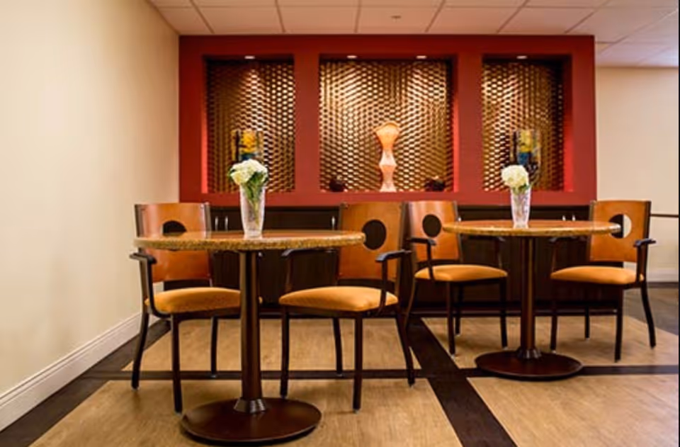 A small dining area with two round tables, each surrounded by three wooden chairs with orange cushions. Each table has a glass vase with white flowers. The background features a decorative wall with a red frame and textured panels, along with artistic sculptures displayed in recessed sections.