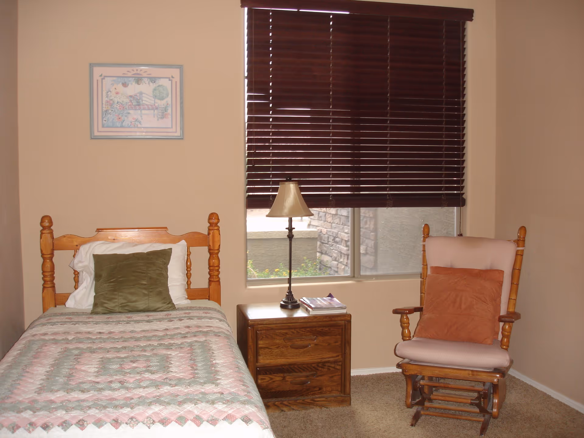 A small bedroom with a single wooden bed covered with a pastel-colored quilt and a green pillow. Next to the bed is a wooden nightstand with a lamp and some magazines. A wooden rocking chair with a pink cushion and an orange pillow is placed near the window, which has dark brown blinds partially closed. A framed picture hangs on the beige wall above the bed.