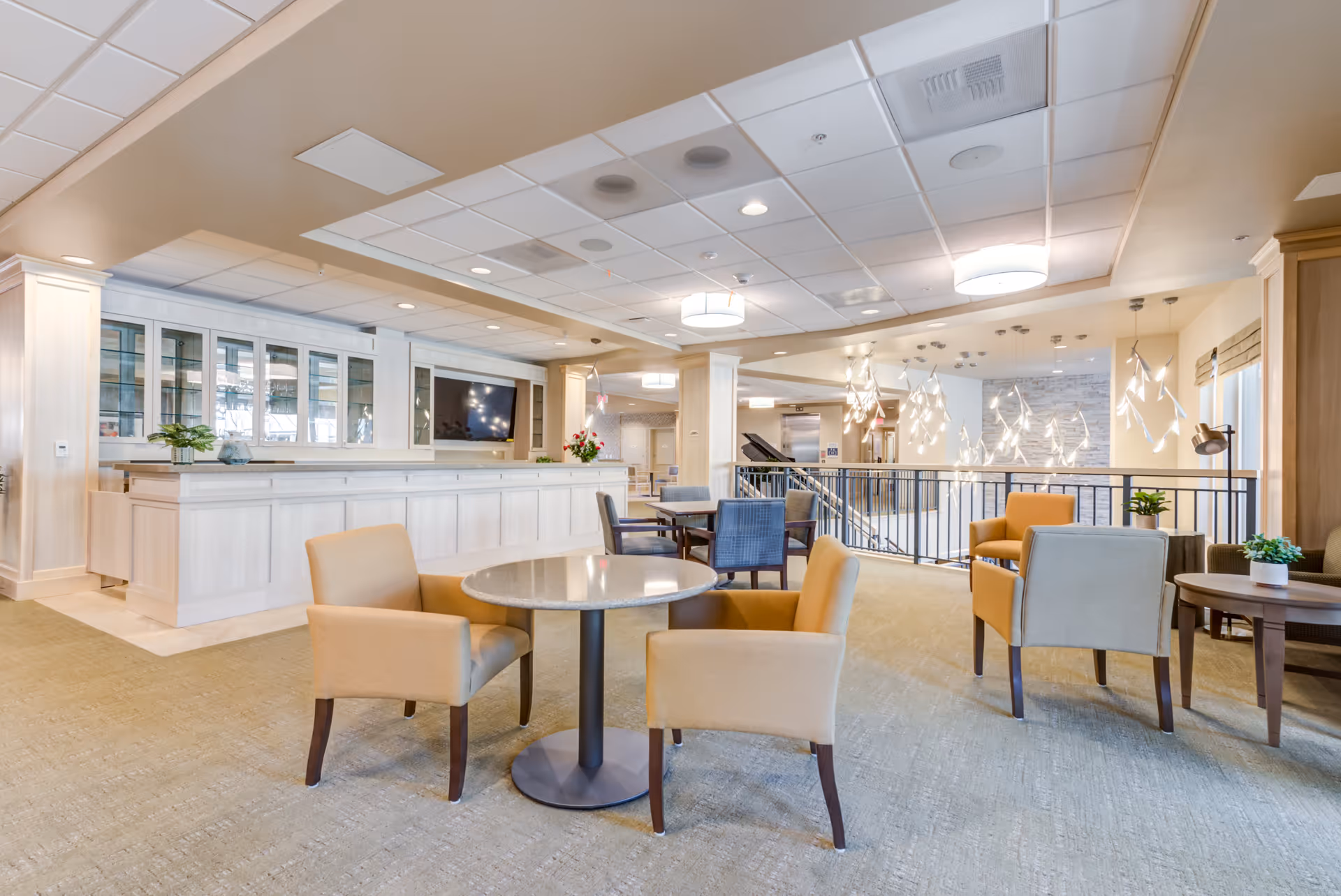 A bright and spacious common area in a senior living facility featuring multiple seating arrangements with beige and gray upholstered chairs around small round tables. The area has a light-colored carpet, a white paneled counter with glass cabinets and a mounted flat-screen TV. Modern pendant lights hang from the ceiling near a railing overlooking a lower level.
