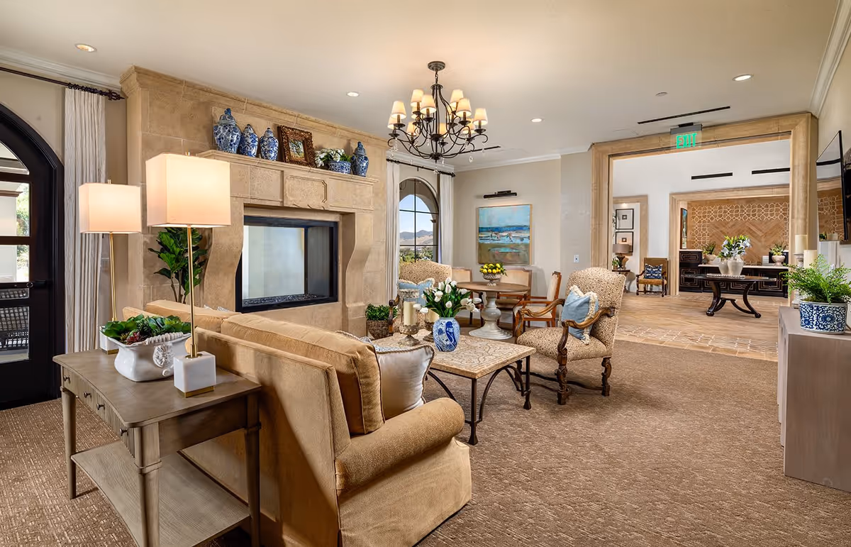 A cozy living room area with beige carpet and furniture including a sofa and armchairs. There is a stone fireplace adorned with blue and white vases and framed pictures. A chandelier hangs from the ceiling, and a table with a vase of flowers is in the center. Large windows and an open doorway lead to another room with additional seating and decorative plants.