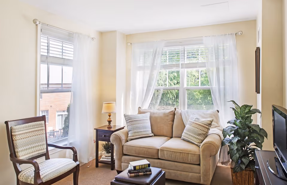 Bright living room with a beige sofa, patterned armchair, side table with lamp, TV and large windows with sheer curtains.