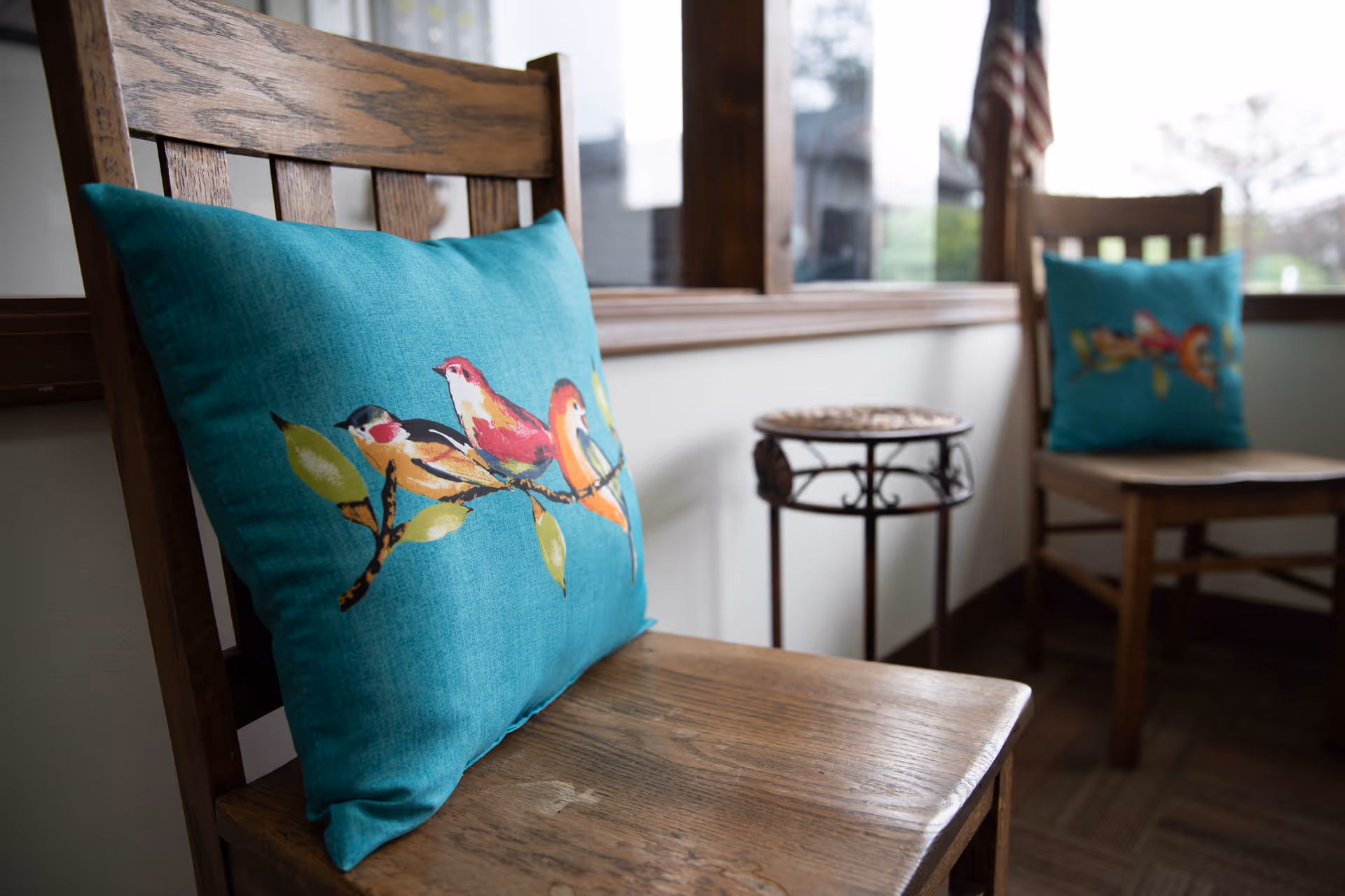 Two wooden chairs with teal cushions featuring colorful bird designs placed near a window with an American flag visible outside.