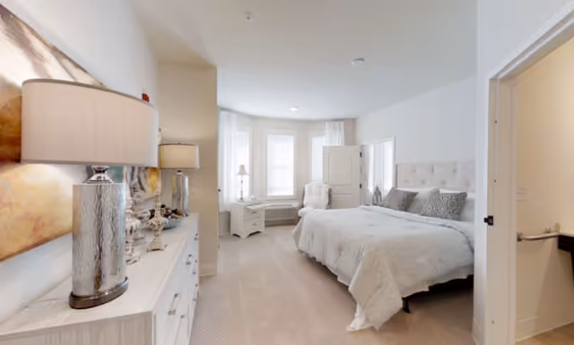 A bright and spacious bedroom with a large bed covered in white bedding, a white dresser with two silver lamps and decorative items, a white armchair near a bay window with white curtains, and a door leading to an adjacent room.