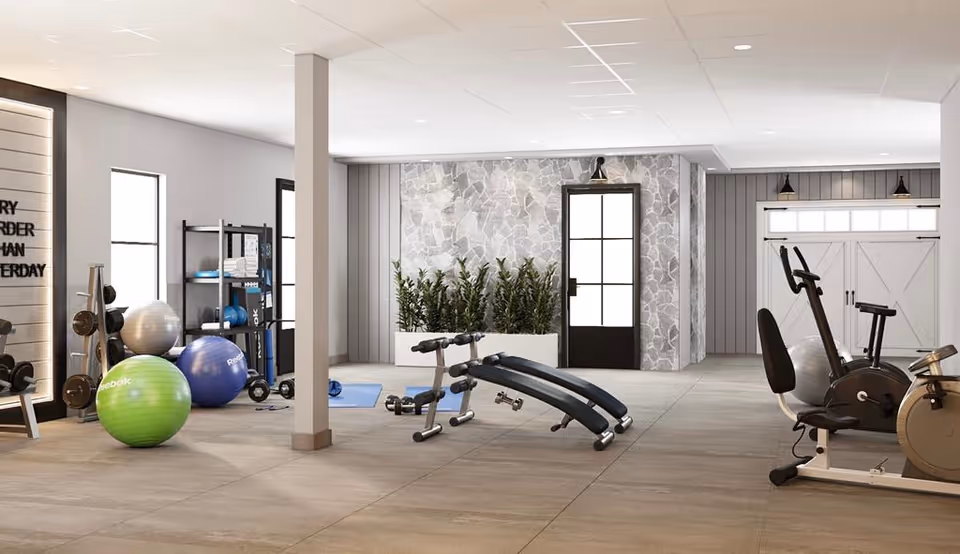 A spacious and well-lit fitness room with exercise equipment including stationary bikes, an adjustable bench, dumbbells, yoga mats, and large exercise balls. The room has large windows, a stone accent wall with a door, and potted plants for decoration.