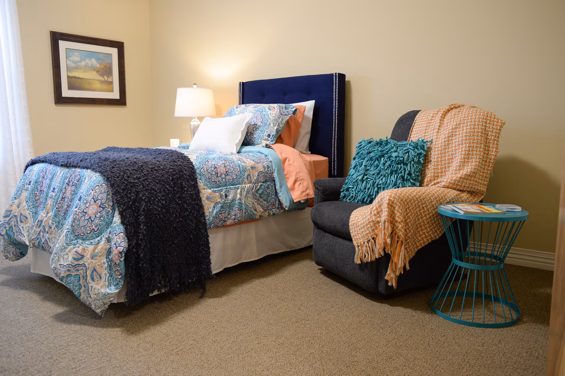 A cozy bedroom featuring a single bed with a blue patterned comforter, white and coral sheets, and a dark blue headboard. Next to the bed is a dark gray armchair draped with an orange throw blanket and a teal textured pillow. A small teal wireframe side table holds some magazines. A lamp on a nightstand and a framed landscape painting adorn the beige walls.