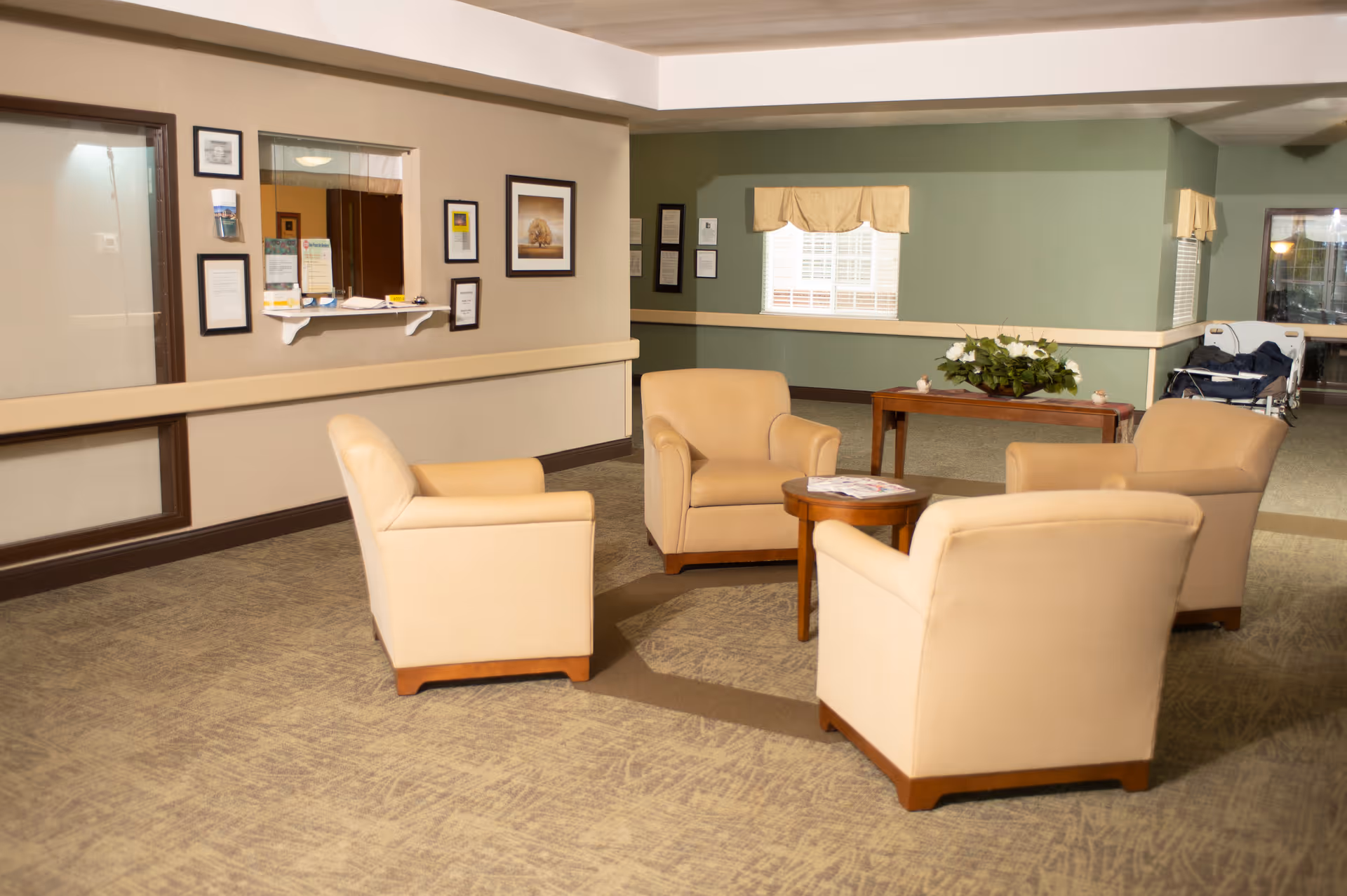 A cozy seating area in a senior living facility with four beige armchairs arranged around a small wooden round table. The room has carpeted floors, light green and beige walls, framed pictures, and a window with a yellow valance. There is a reception window with documents and a hand sanitizer dispenser on the wall, and a table with a plant in the background.