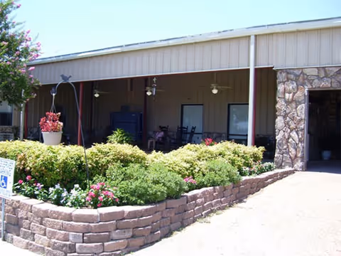 Outdoor covered patio area with ceiling fans and rocking chairs, adjacent to a building with stone and metal siding. In front of the patio is a raised garden bed with various green shrubs and flowering plants. A tree with pink flowers is visible on the left side.