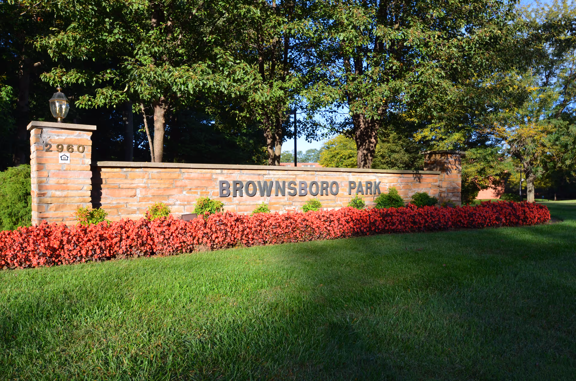 A brick sign for Brownsboro Park Retirement Community surrounded by red flowers and green grass, with trees in the background under a clear blue sky.