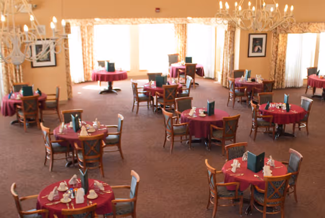 A dining room with multiple round tables covered with red tablecloths, each set with napkins, cups, and menus. Wooden chairs with cushioned seats surround the tables. The room has large windows with patterned curtains and chandeliers hanging from the ceiling.