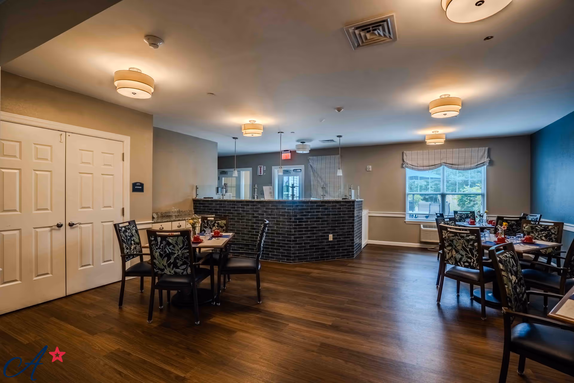 A dining area in an assisted living facility with several tables and chairs arranged neatly. The tables are set with red cups and small flower vases. The room has wooden flooring, beige walls with a blue accent wall, and ceiling lights. There is a window with a checkered valance allowing natural light to enter. A counter with a dark brick facade is visible in the background.