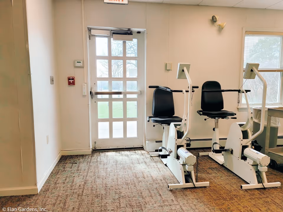 A small exercise room with two white and black stationary recumbent exercise bikes positioned near a glass door and a window. The room has beige carpet flooring and white walls, with a fire alarm and thermostat mounted on the wall near the door.