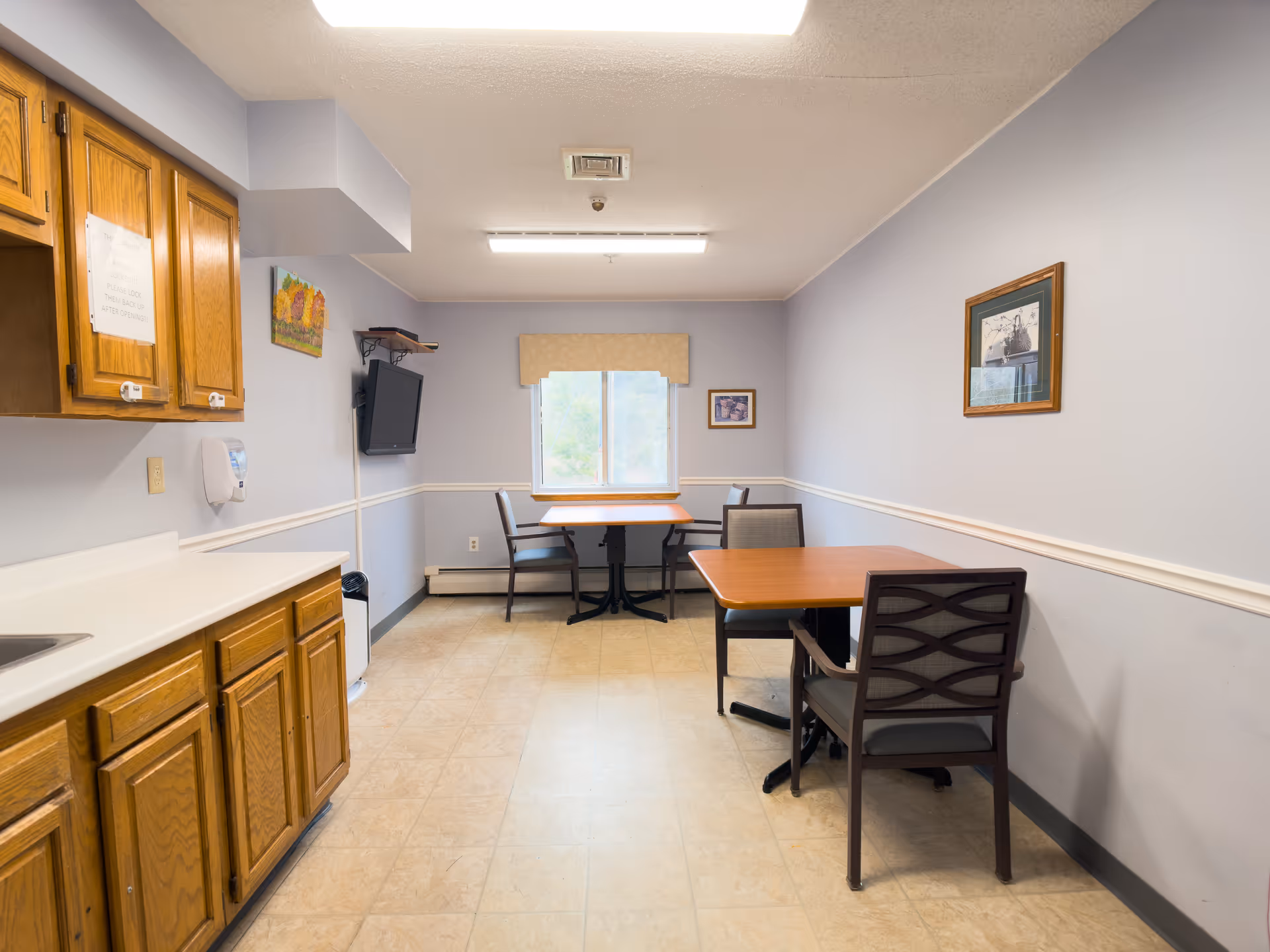 A small dining area in a senior living facility with two wooden tables and four chairs. There is a window with a beige valance at the far end of the room, light blue walls with white trim, and a wall-mounted television. On the left side, there are wooden cabinets and a countertop with a sink. The floor is tiled, and there are framed pictures on the walls.