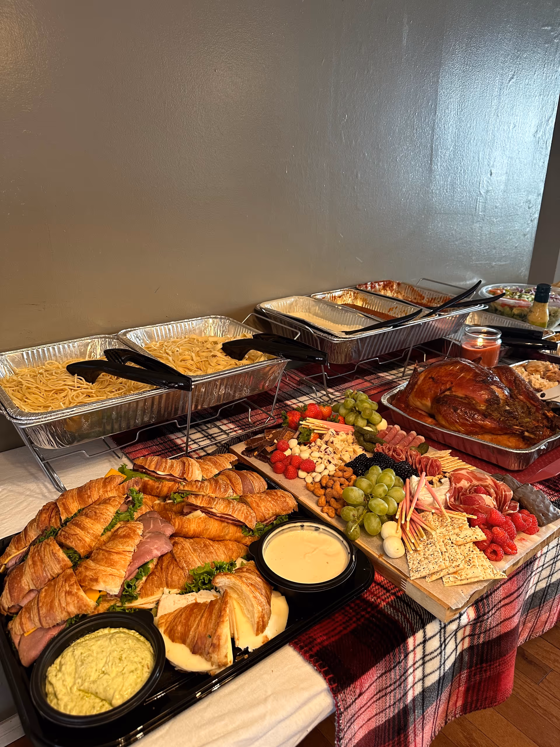 A buffet-style spread featuring trays of pasta, a roasted turkey, a charcuterie board with grapes, berries, nuts, cheese, crackers, and sliced meats, as well as croissant sandwiches with lettuce and deli meat, all arranged on a table covered with a plaid tablecloth.