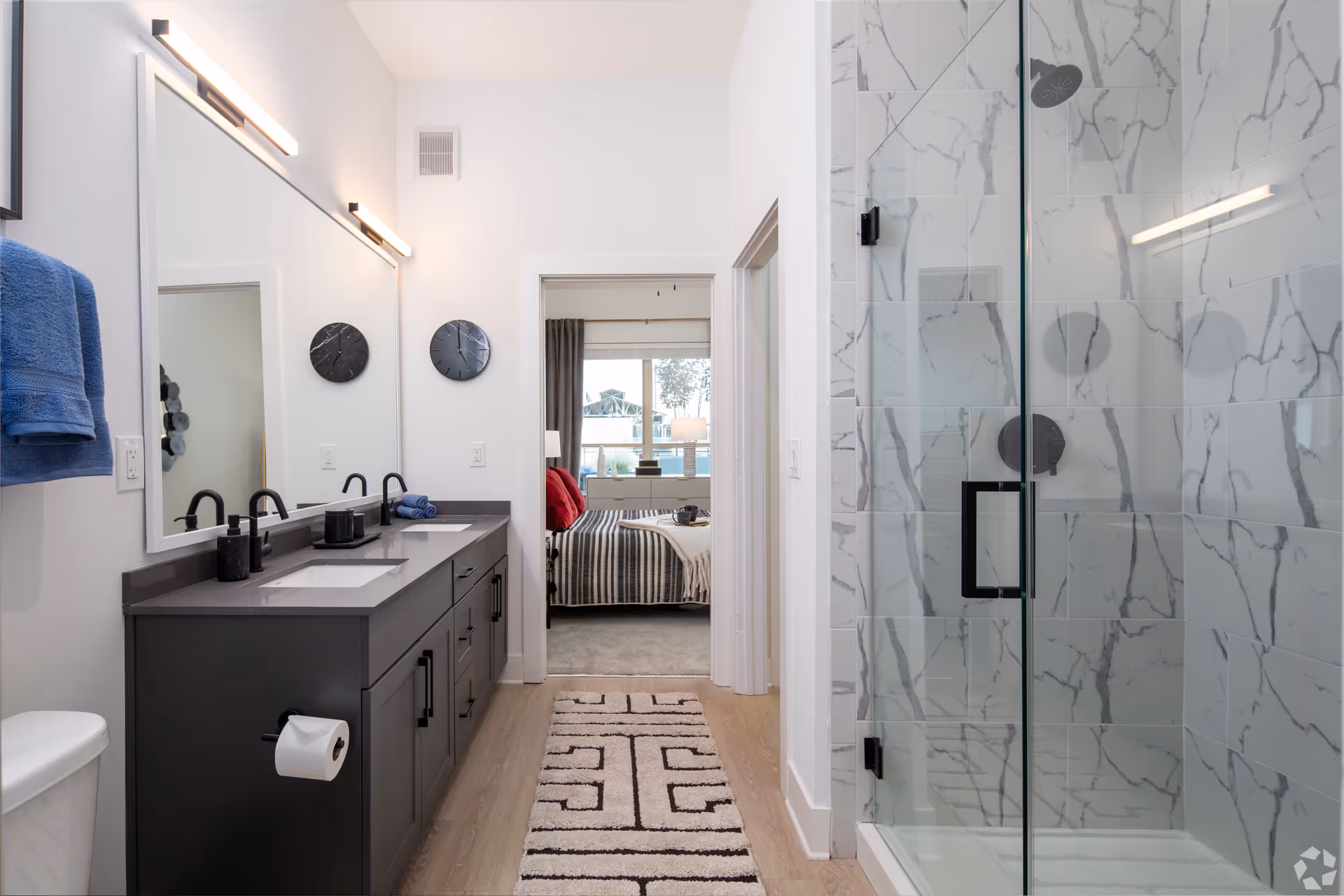 Modern bathroom with a double sink vanity featuring black faucets and a large mirror above. A glass-enclosed shower with white marble-patterned tiles is on the right. A blue towel hangs on the left wall, and a patterned rug runs along the wooden floor. Through the doorway, a bedroom with a striped bedspread and red pillows is visible.