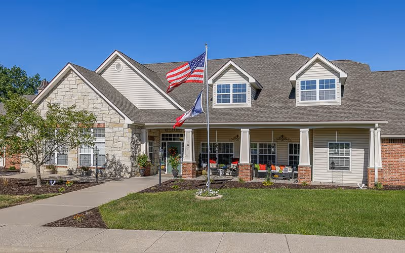 Front exterior of a single-story senior living building with a covered porch, flagpoles, and a manicured lawn.