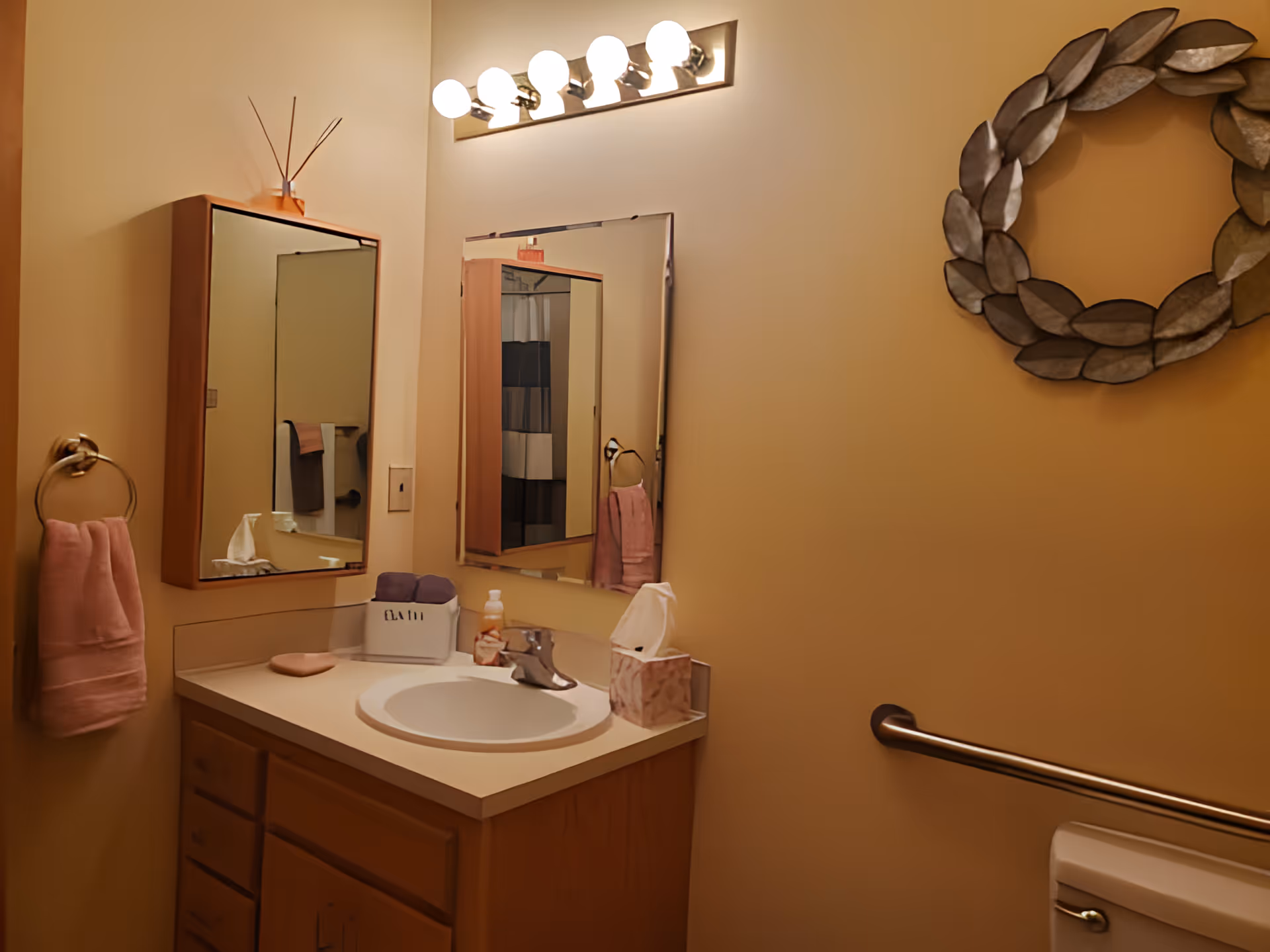Small bathroom with a sink vanity and mirror, pink towels, decorative wall wreath, and a grab bar by the toilet.
