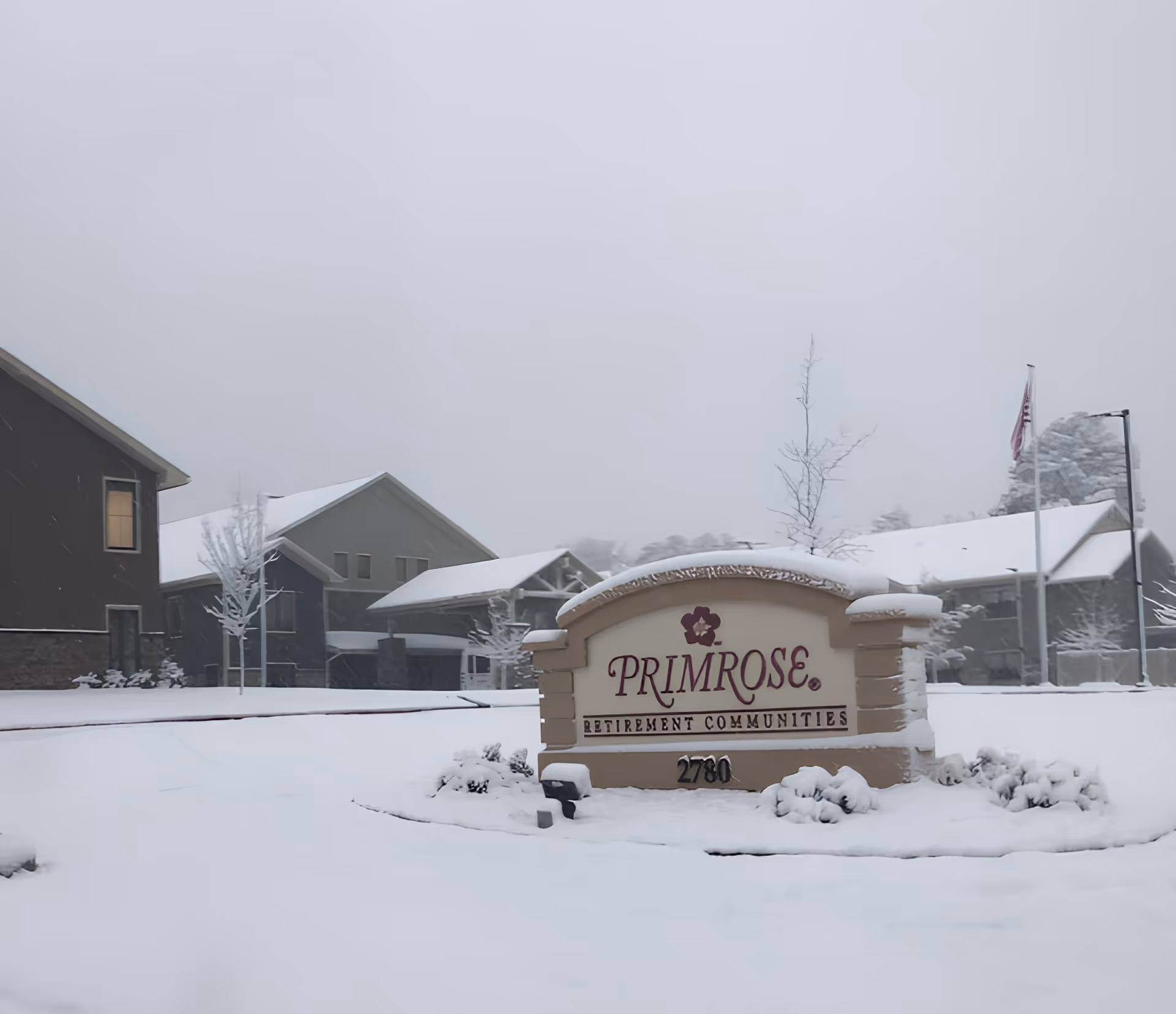 Snow-covered entrance sign reading 'Primrose' in front of a retirement community building and grounds.