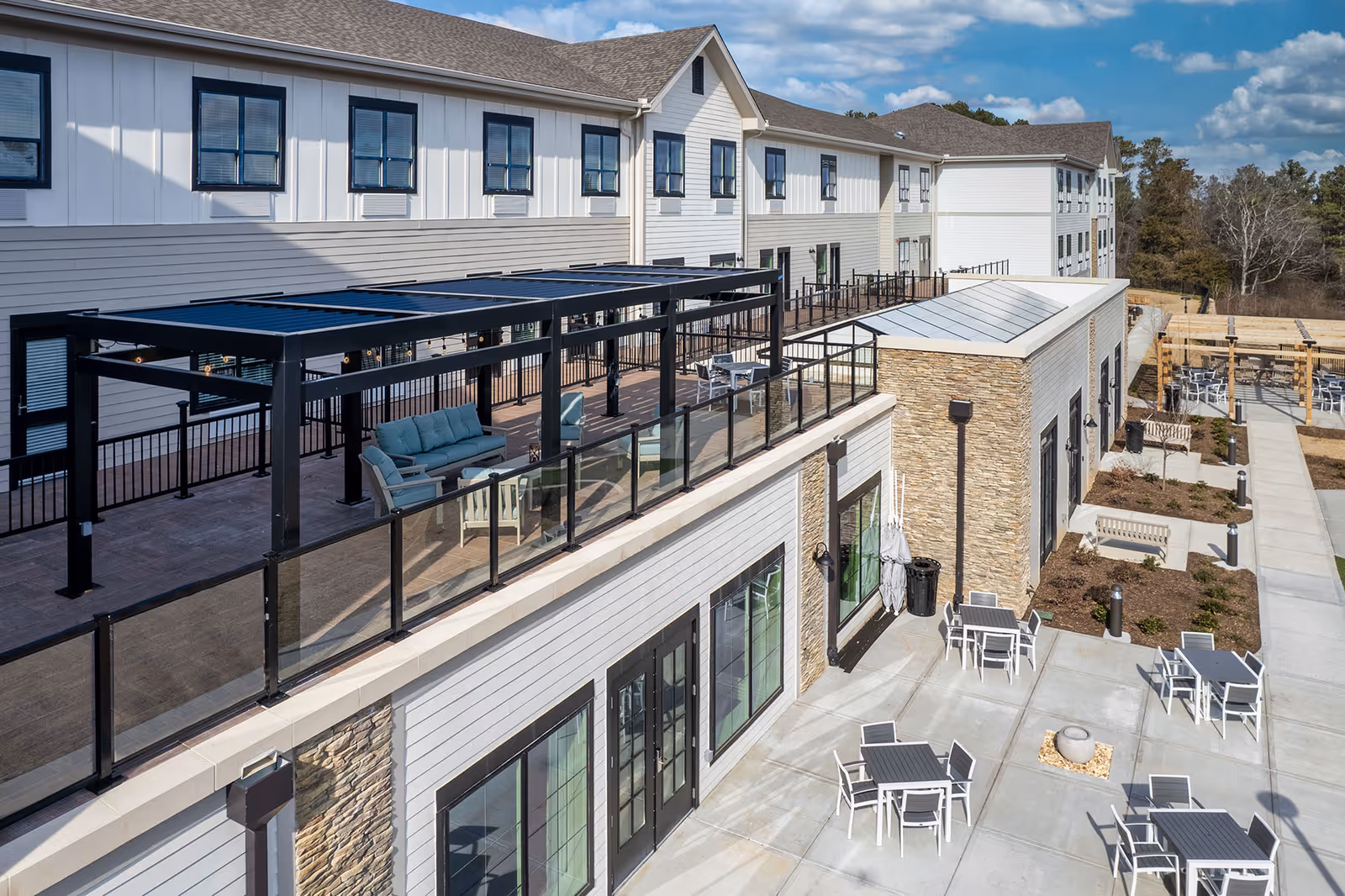 Outdoor patio area of Cloverland Park Senior Living featuring multiple seating arrangements with tables and chairs on a concrete surface. The building has a modern design with white siding, stone accents, and large windows. There is a covered seating area with a black pergola and cushioned chairs on an elevated deck. Trees and a partly cloudy sky are visible in the background.