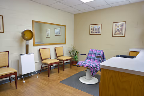Small salon-style room with a styling chair covered by a plaid cloth, hooded hair dryer, waiting chairs, mirror, and countertop.