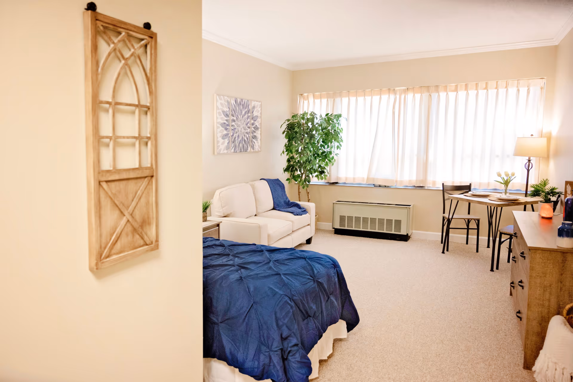 A cozy, well-lit bedroom with a bed covered in a dark blue comforter, a white loveseat with a blue throw blanket, a small dining table with two chairs near a large window with sheer curtains, a wooden dresser with decorative items, a floor lamp, and a green potted plant in the corner.