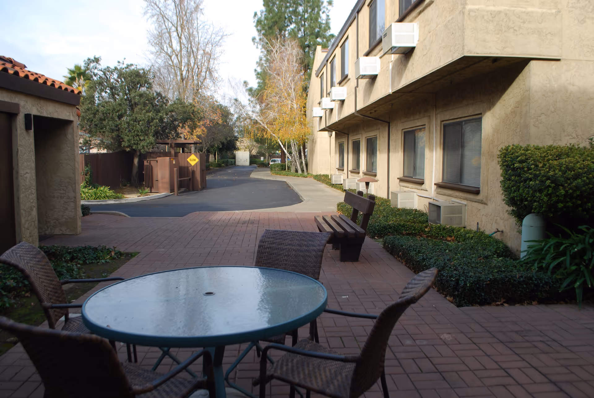 Outdoor patio area at Leisure Manor Apartments with a round glass table and four wicker chairs in the foreground. There is a wooden bench along the side of a beige building with multiple windows and air conditioning units. Trees and bushes surround the paved walkway and driveway in the background.