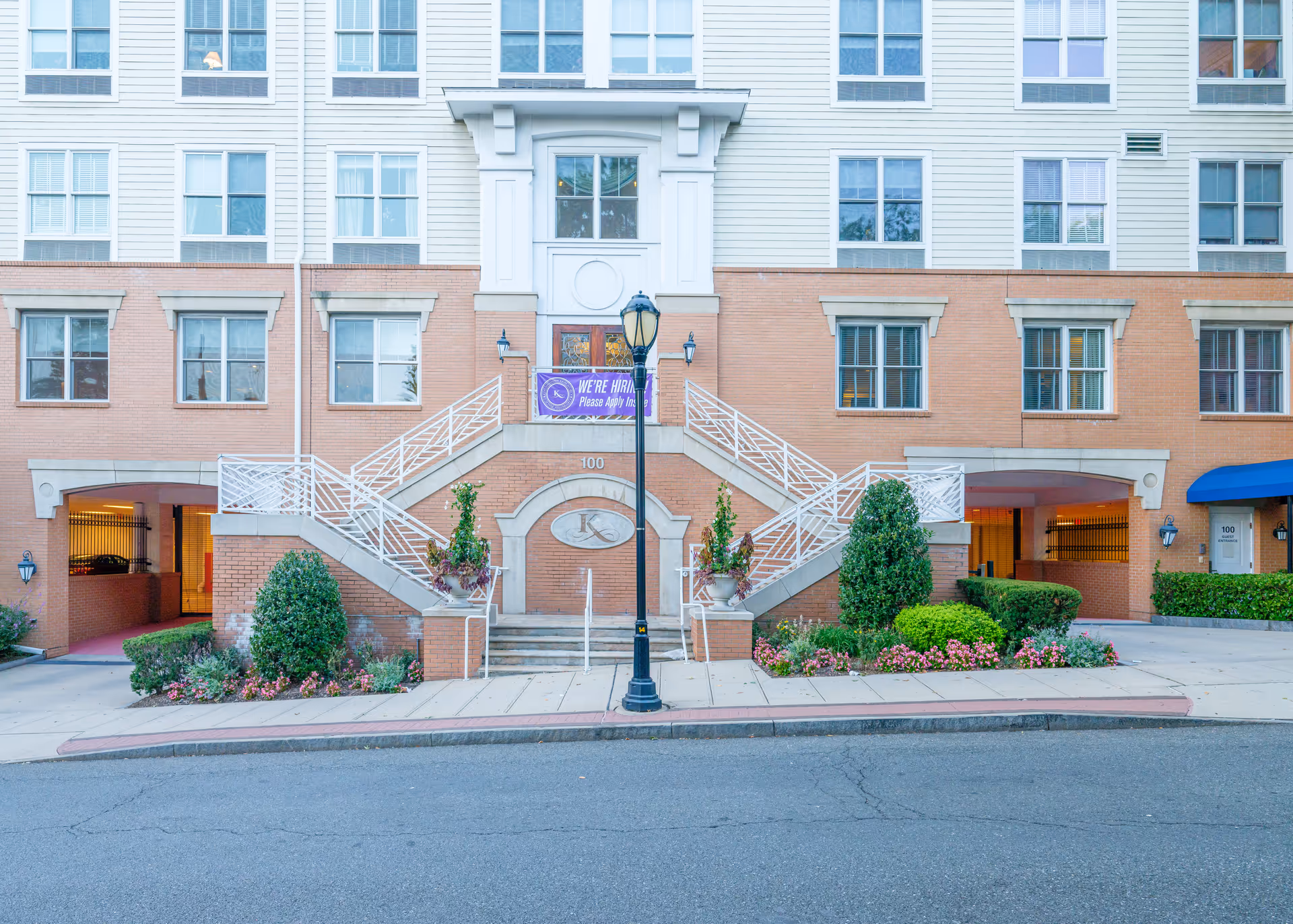 Front facade of a multi-story brick-and-siding senior living building with symmetrical exterior staircases, landscaped planters and a central entrance.