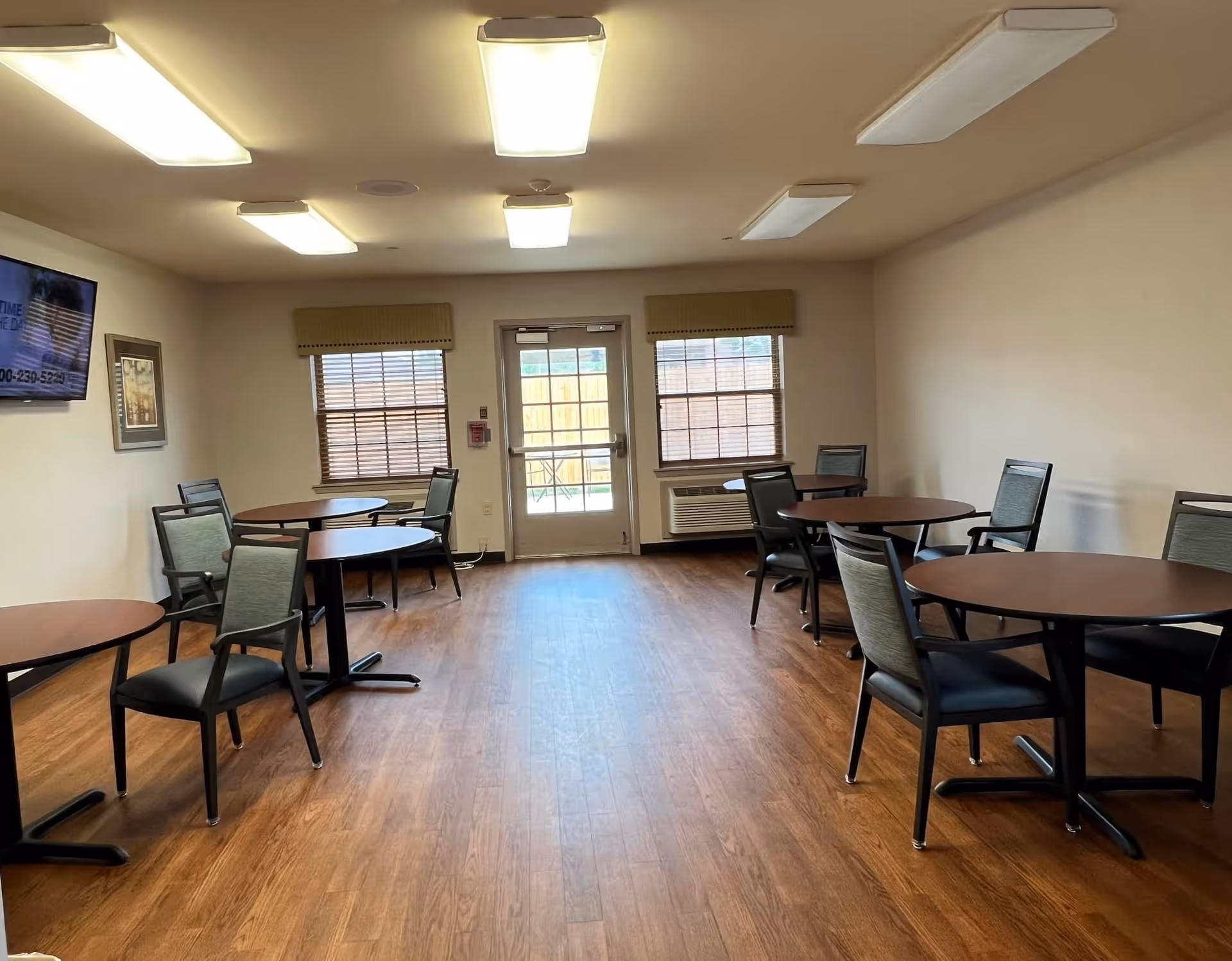 Spacious common dining/activity room with round tables and chairs, wood flooring, ceiling lights, windows and a door to the outside.
