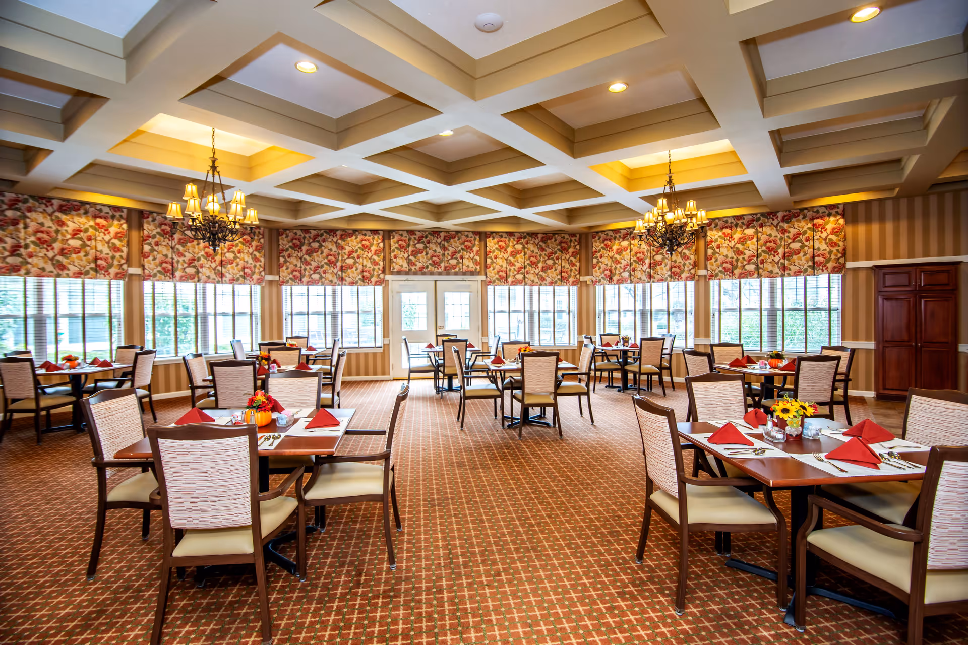 A spacious dining room with multiple tables set with red napkins, white placemats, and small floral centerpieces. The room features large windows with floral-patterned valances, a coffered ceiling with recessed lighting, and two ornate chandeliers. The carpet has a geometric pattern, and the walls are decorated with striped wallpaper and wooden accents.