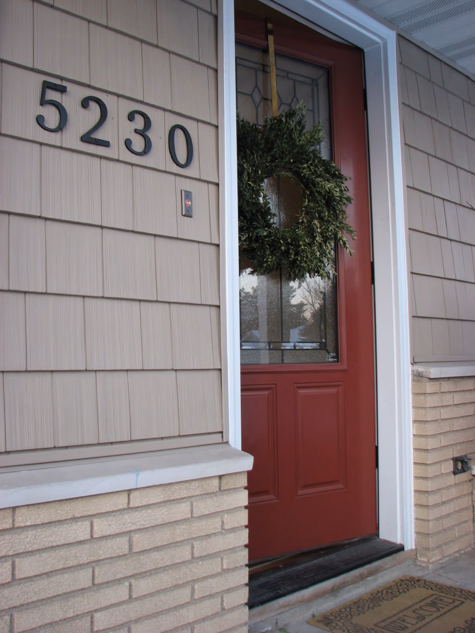 Front entrance of a building with a red door decorated with a green wreath. The building has beige siding and brickwork below the door. The house number 5230 is displayed on the wall next to the door, and a welcome mat is placed on the doorstep.