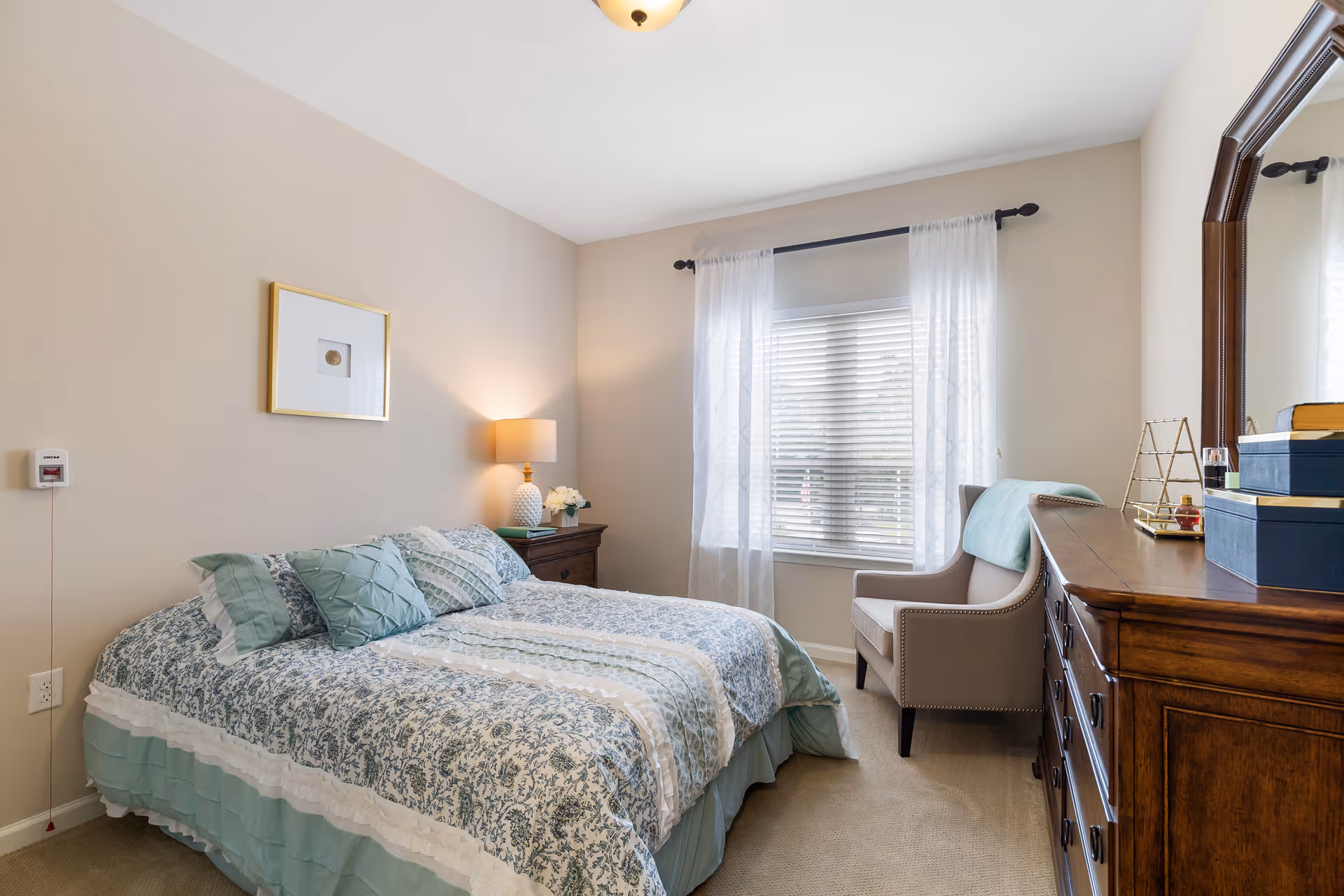 Well-lit bedroom with a made bed, bedside table and lamp, dresser with mirror, and an armchair by a window with sheer curtains.
