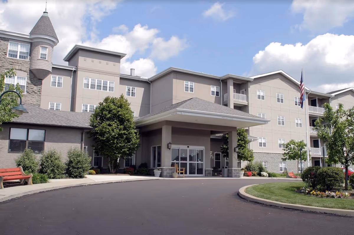 Front entrance of a multi-story senior living facility with a covered porte-cochere, landscaping, benches, and an American flag.
