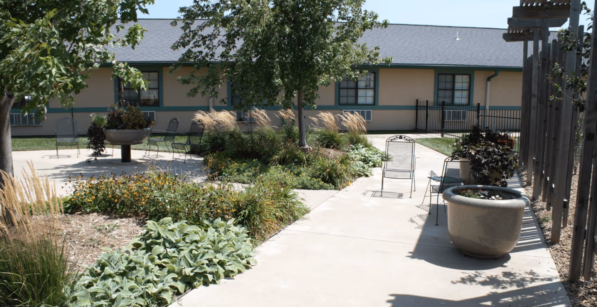 Outdoor garden area with paved walkways, metal chairs, various plants, trees, and large planters, adjacent to a single-story building with beige walls and green trim.