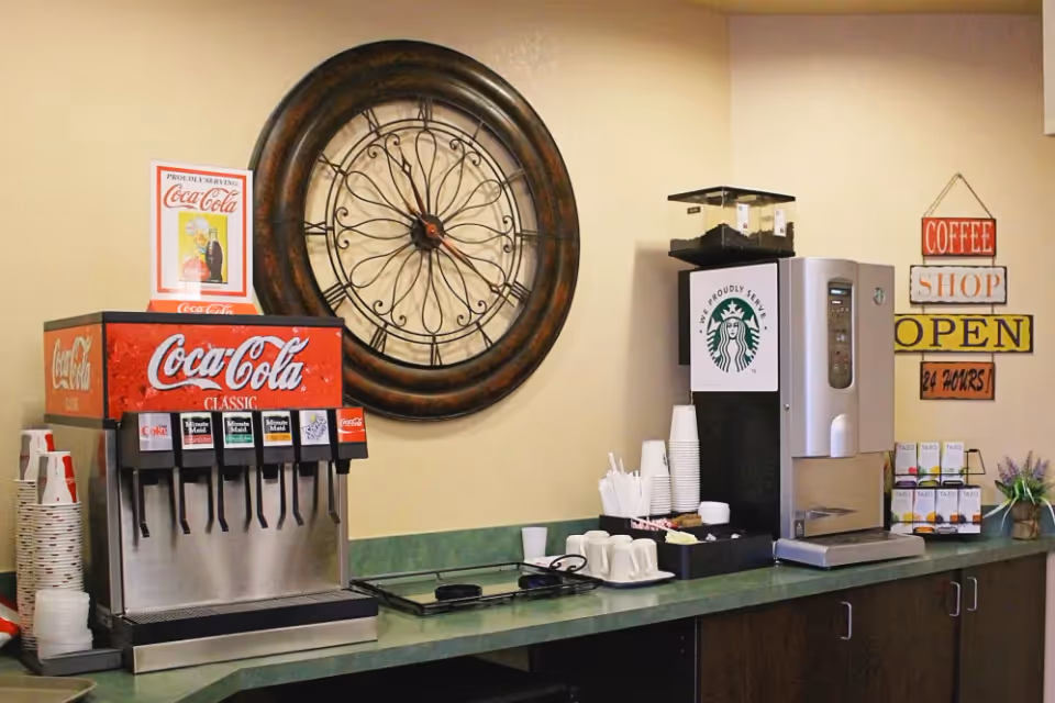 A beverage station with a Coca-Cola soda fountain machine and a Starbucks coffee dispenser on a green countertop. There are cups, lids, stirrers, and various tea boxes arranged neatly. A large decorative wall clock and signs reading 'Coffee Shop Open 24 Hours' and 'Proudly Serving Coca-Cola' are visible on the beige wall behind the station.