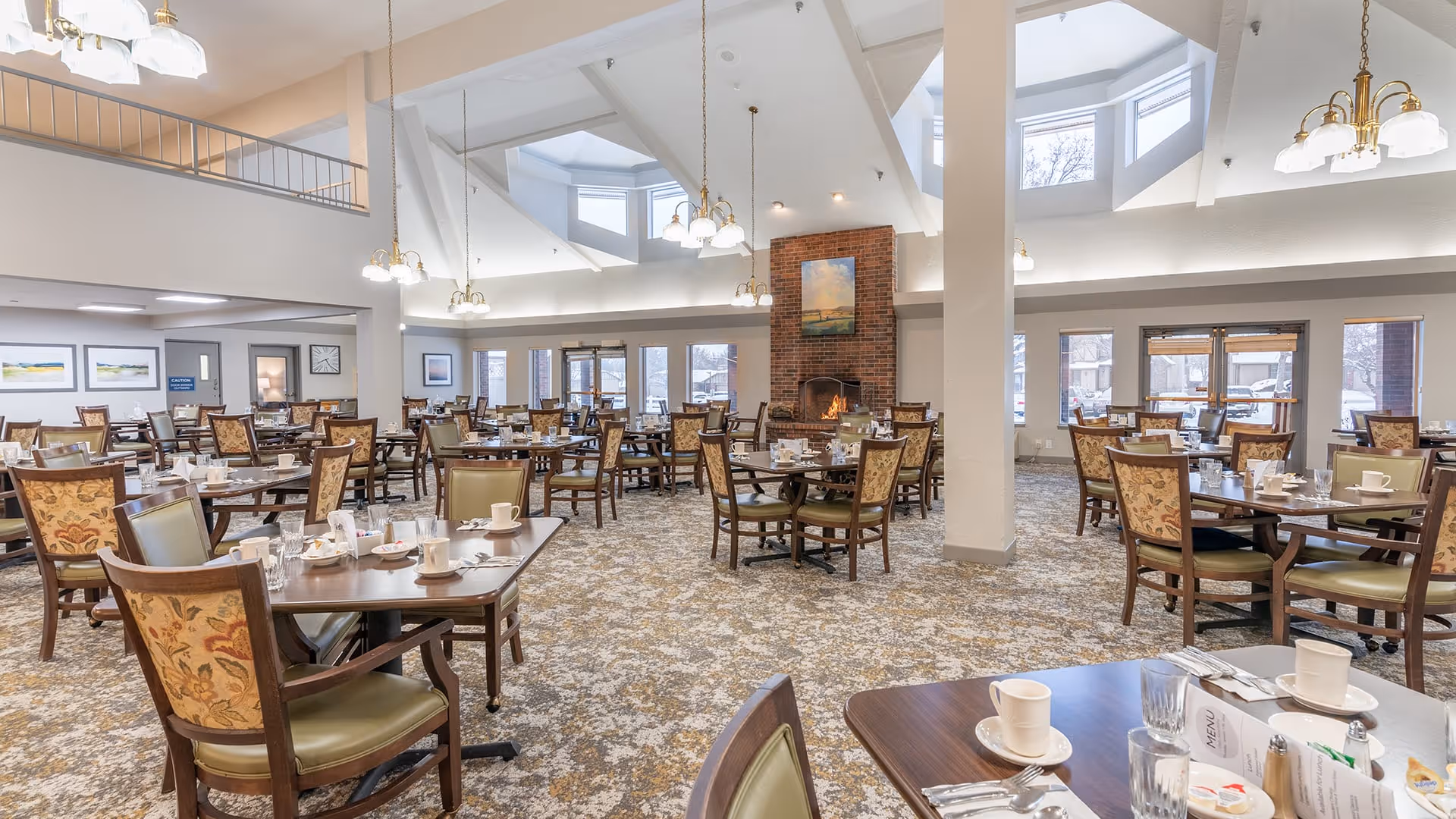 A spacious dining room with multiple wooden tables and chairs arranged neatly. Each table is set with white cups, glasses, napkins, and silverware. The room features a high ceiling with skylights, hanging light fixtures, and a brick fireplace with a painting above it. Large windows allow natural light to enter the room.