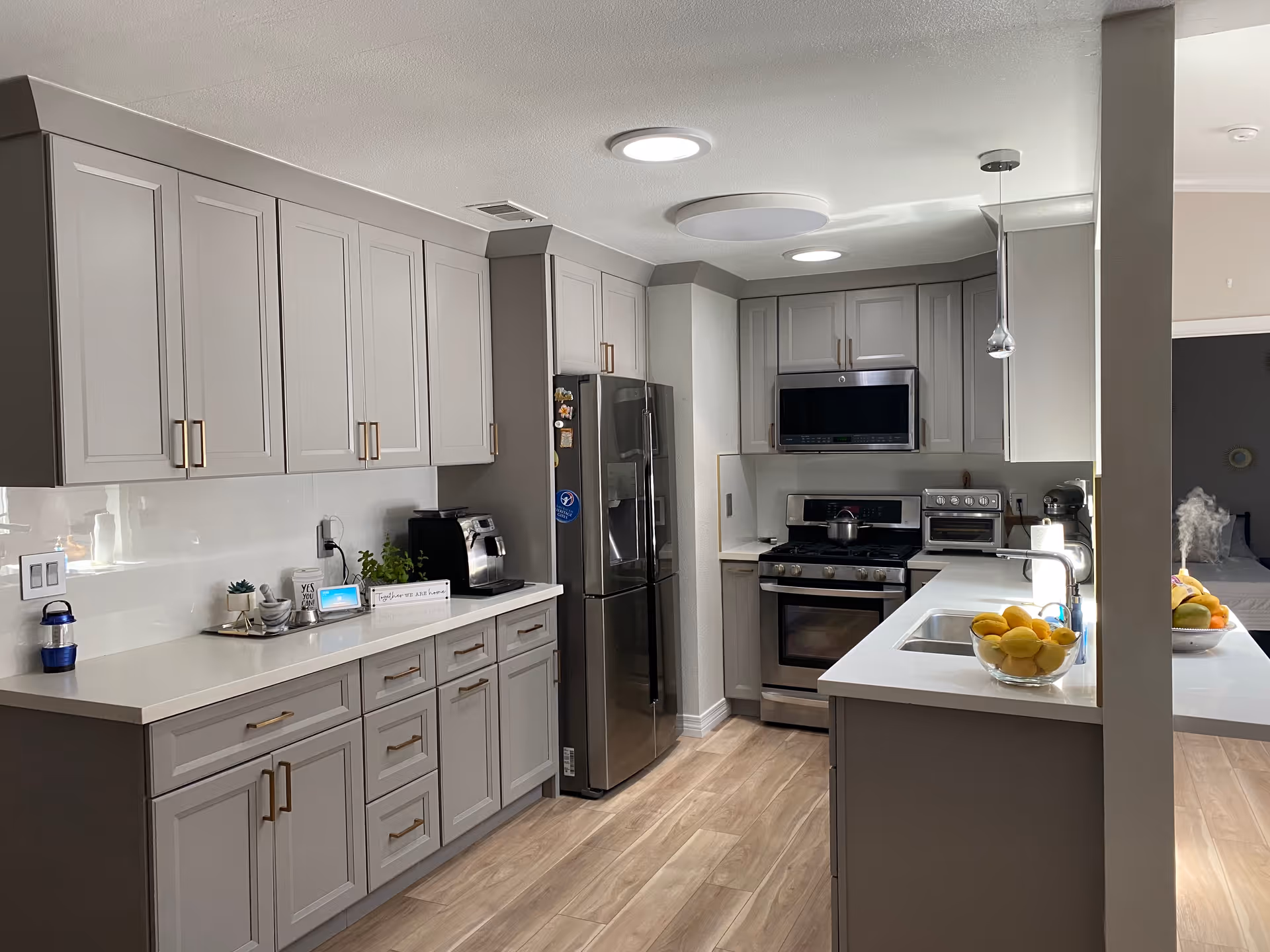 Modern kitchen with light gray cabinets, stainless steel refrigerator, oven, microwave, and various kitchen appliances on the countertops. The kitchen has wood flooring and a white countertop with a sink and a bowl of lemons. In the background, a bedroom is partially visible.