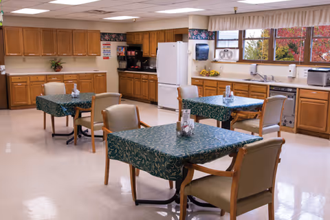 A bright and clean dining area with three tables covered in green patterned tablecloths, each surrounded by four beige cushioned chairs. The room features wooden cabinets along the walls, a white refrigerator, a dishwasher, a microwave, and a coffee machine. Large windows let in natural light and show colorful autumn foliage outside.