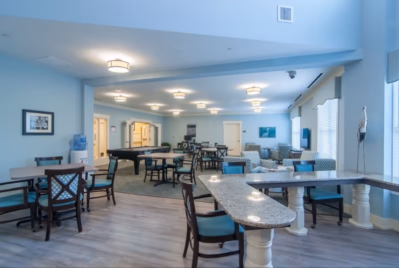 A spacious common area in a senior living facility with multiple tables and chairs, a pool table, comfortable armchairs, and large windows letting in natural light. The room has light blue walls, modern ceiling lights, and a water cooler in the corner.