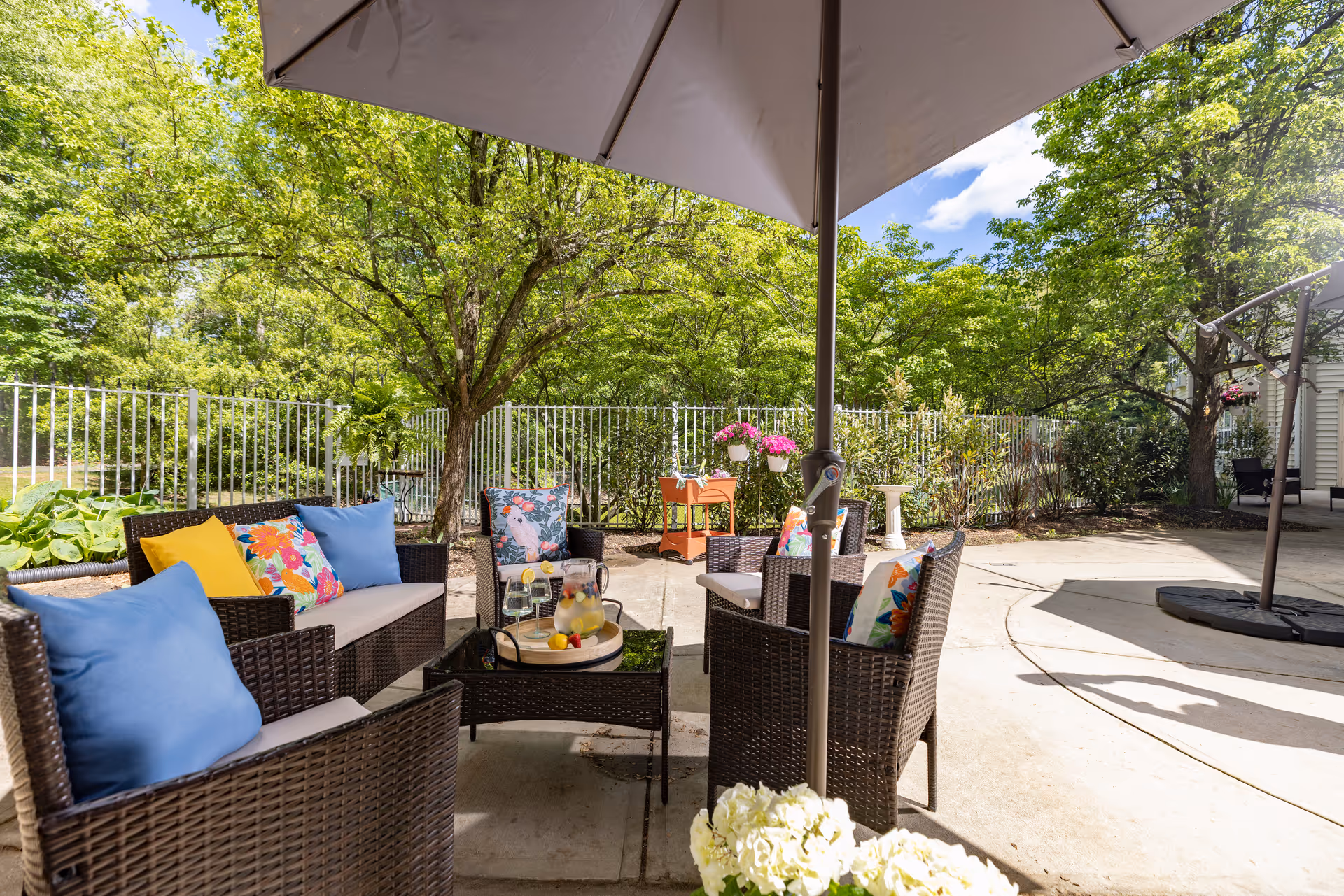 Outdoor patio area with wicker furniture including chairs and a sofa adorned with colorful cushions. A large umbrella provides shade over a glass-top table with a pitcher of lemonade and glasses. The patio is surrounded by green trees and plants, with a white fence in the background under a blue sky with some clouds.