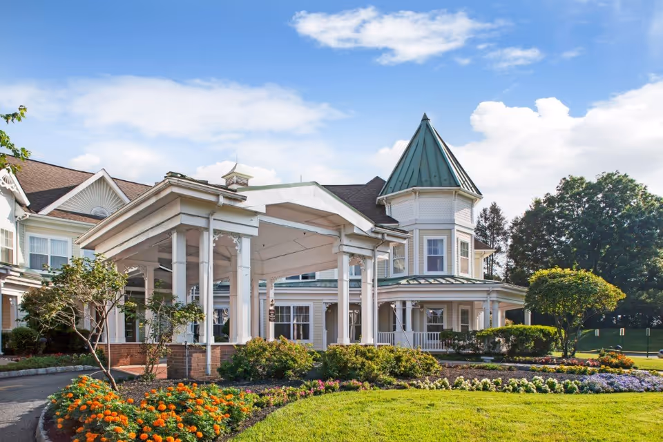 Exterior view of Sunrise of Westfield facility featuring a large covered entrance with white columns, a turret with a green roof, well-maintained flower beds with orange and purple flowers, green shrubs, and a manicured lawn under a partly cloudy blue sky.