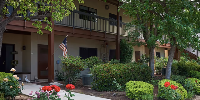Exterior of a two-story retirement community building showing ground-floor entrances, an upper balcony, landscaped shrubs and red roses.