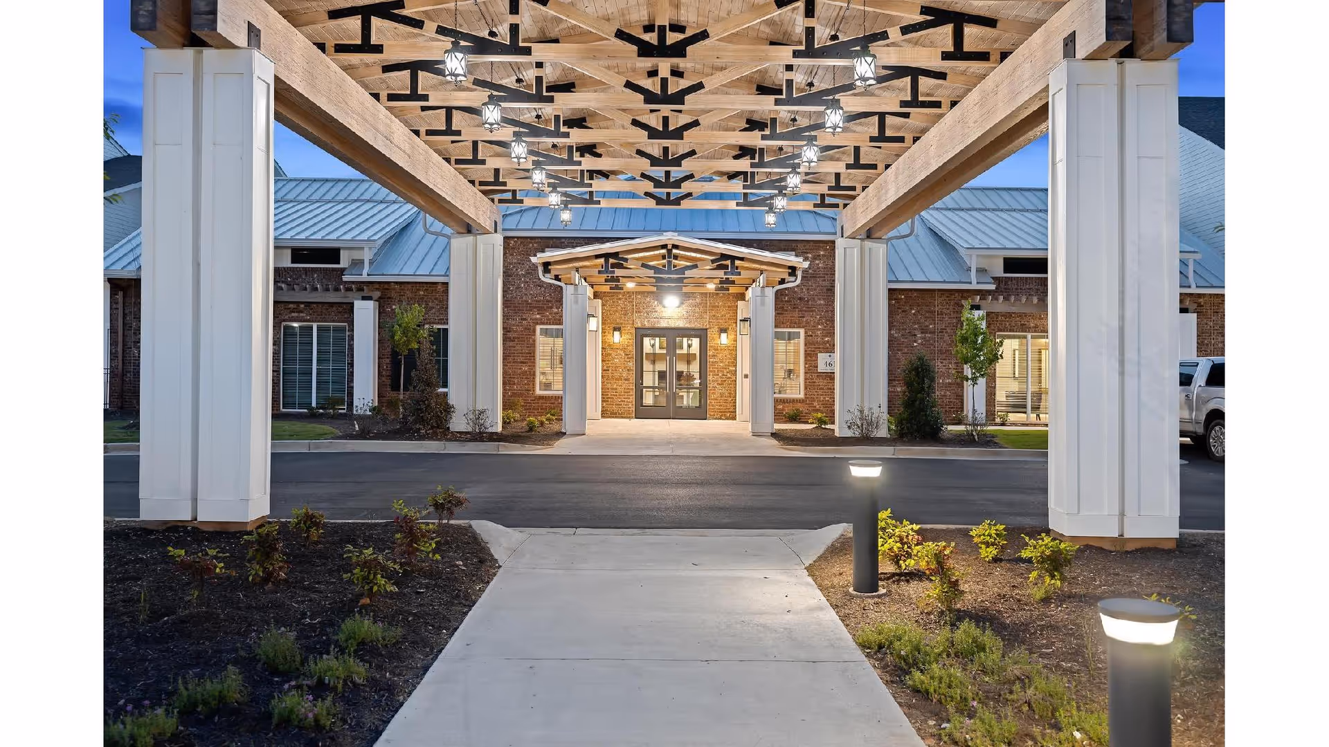 Entrance to a senior living facility with a covered driveway supported by large white columns and wooden beams with decorative lighting. The building has brick walls and a metal roof, with a concrete walkway leading to the glass double doors entrance. Small landscaped areas with plants and illuminated bollard lights flank the walkway.