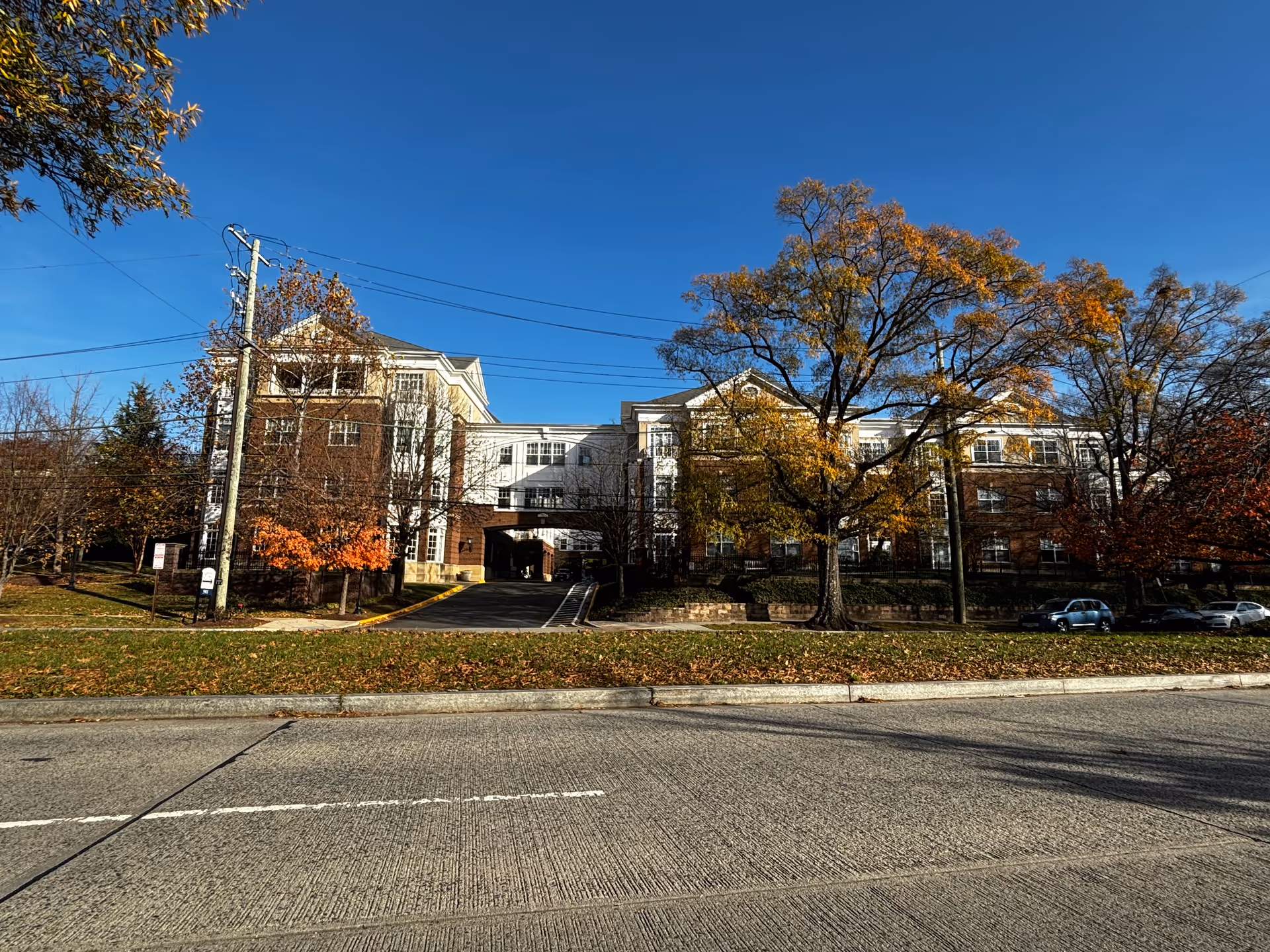 Exterior view of a multi-story assisted living community building with a combination of brick and white siding. The building has large windows and an archway driveway entrance. Trees with autumn foliage surround the building under a clear blue sky.