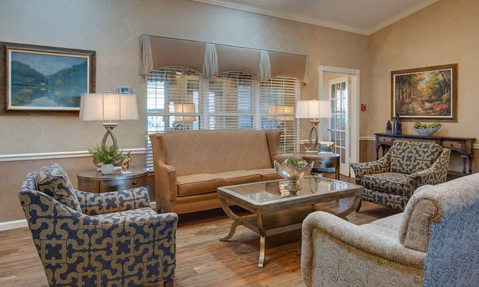 A cozy senior living facility lounge with a tan leather sofa, two patterned armchairs, and a beige armchair arranged around a wooden coffee table with a decorative bowl. The room features wooden flooring, two side tables with lamps, framed landscape paintings on the walls, and large windows with blinds and valances.